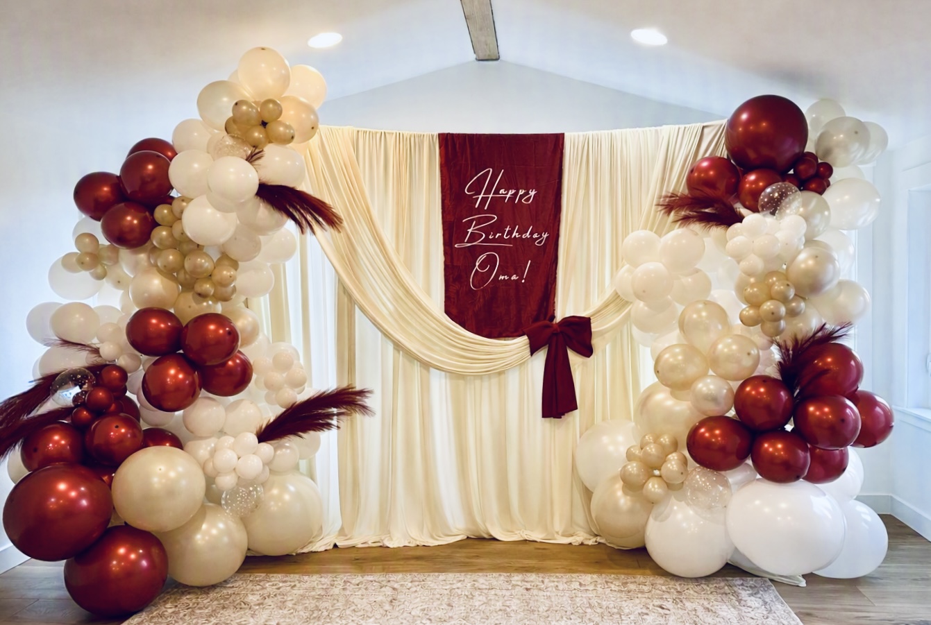 Burgundy and cream balloon arch with pampas grass around a draped birthday backdrop